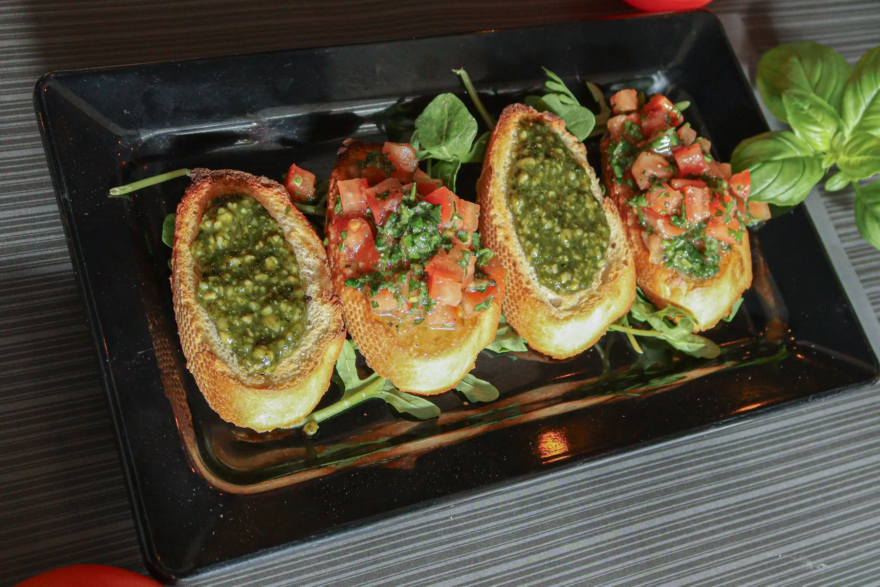Bruschetta: vier sneetjes brood met tomaat, pesto en basilicum op een zwarte plank